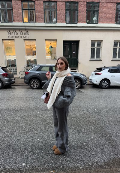 Suéter gris de gran tamaño con diseño off-the-shoulder, bufanda de punto color crema, pantalones grises y botas marrones. Calle peatonal con coches aparcados.