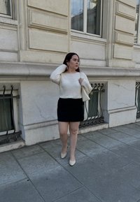 Pull en tricot blanc, jupe mini noire et ballerines à bout pointu beiges, accompagnés d'un petit sac à main blanc. Vue de profil contre un bâtiment en pierre.