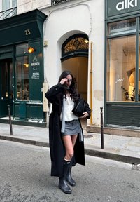 Žena v černém kabátě, šedé sukni a černých botách si upravuje sluneční brýle, stojíc na městské ulici u zeleného obchodu a zaklenutých dveří.