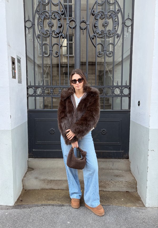 Brown faux fur jacket with a hood, light grey top, wide-leg blue jeans, brown handbag, and brown slip-on shoes, standing in front of a black wrought-iron gate.