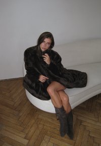 Brun faux pelsfrakke med en blød tekstur, der har brede ærmer. Båret over en siddende figur med lysebrune ben og grå knælange støvler på en buet sofa.