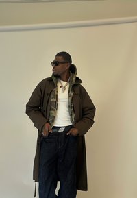 Mann mit dunkler Sonnenbrille, braunem Trenchcoat in Lagen über einer Camouflage-Jacke, weißem Hemd, dunklen Jeans und silbernem Schmuck, der gegen eine beige Wand steht.
