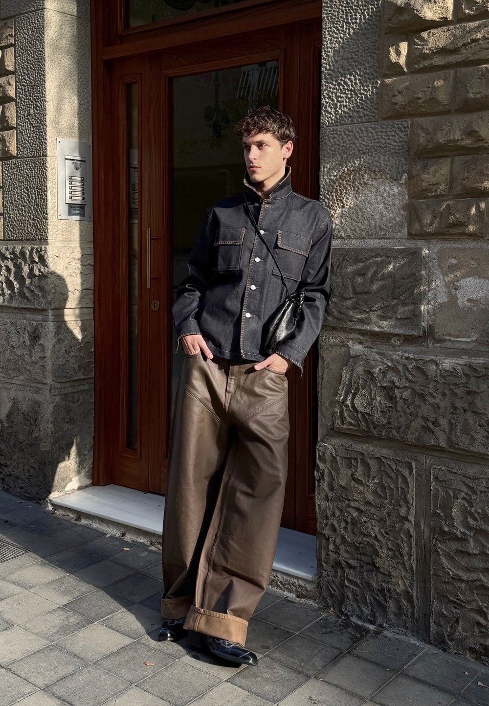Veste en denim noire avec fermeture à boutons et poches avant, associée à un pantalon en cuir beige. Debout contre un mur en pierre. Sac à main noir.