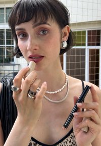 Rouge à lèvres dans un tube beige tenu par une personne avec des bijoux en argent. L'arrière-plan montre un motif en grille. La peau a des taches de rousseur ; les yeux sont marron clair.