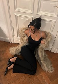 Manteau en fausse fourrure beige clair porté sur un débardeur noir et un pantalon noir taille haute. Talons pointus noirs et sol en bois en arrière-plan.