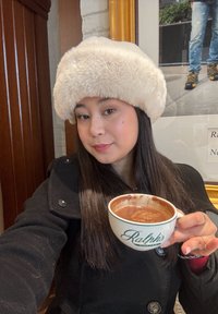 Jonge vrouw met een beige, pluizige hoed en een zwarte jas houdt een kop warme chocolademelk vast in een koffiewinkel.