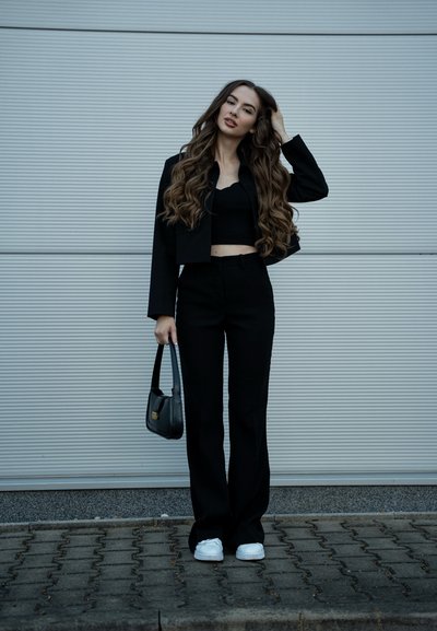 Blazer negro corto sobre un top corto negro con bordes ondulados, combinado con pantalones de campana negros y zapatillas blancas. Sosteniendo un bolso negro.