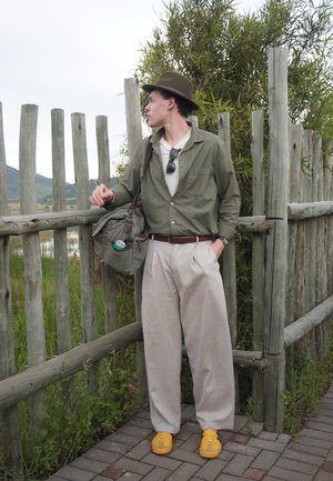 Hombre con camisa verde y sombrero está junto a una valla de madera con una bolsa verde y zapatos amarillos, mirando de lado en un entorno natural al aire libre.