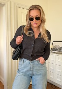 Grå strikket cardigan med knappelukking, lyseblå høytlivsjeans og svart håndveske. Runde solbriller og langt, bølgete hår.
