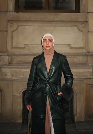 Mujer con hiyab blanco, abrigo largo de cuero verde oscuro con puños de piel, suéter de cuello alto color beige, sosteniendo un bolso y de pie frente a un fondo de pared de piedra.