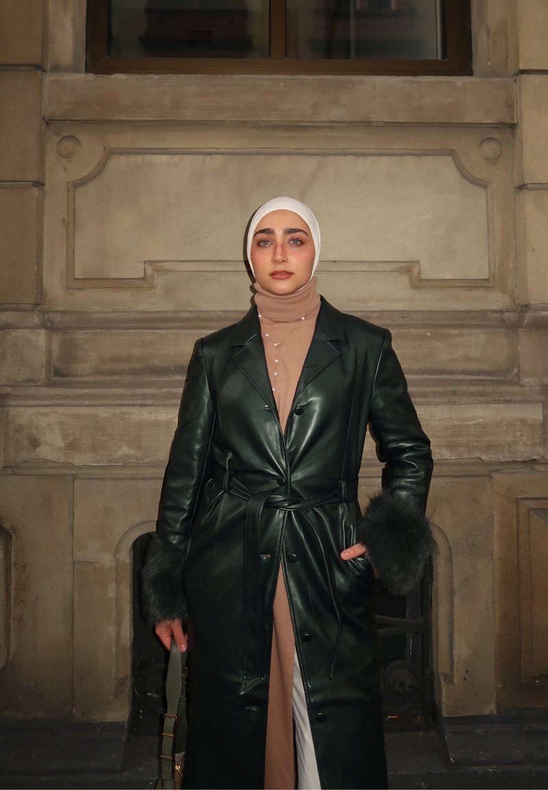 Donna che indossa un hijab bianco, un lungo cappotto di pelle verde scuro con polsini in pelliccia, un dolcevita color caffelatte, tiene una borsa, in piedi contro uno sfondo di parete di pietra.