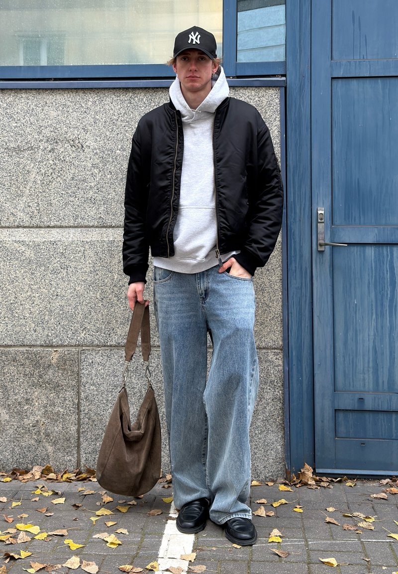 Giubbotto bomber nero sopra una felpa con cappuccio grigia, jeans larghi azzurri e scarpe nere. Borsa marrone con tracolla. Ambientazione urbana con parete testurizzata.