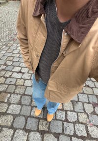 Veste légère couleur beige avec un col en velours côtelé marron, portée sur un pull gris côtelé et un jean bleu clair, assortie à des sabots jaunes.