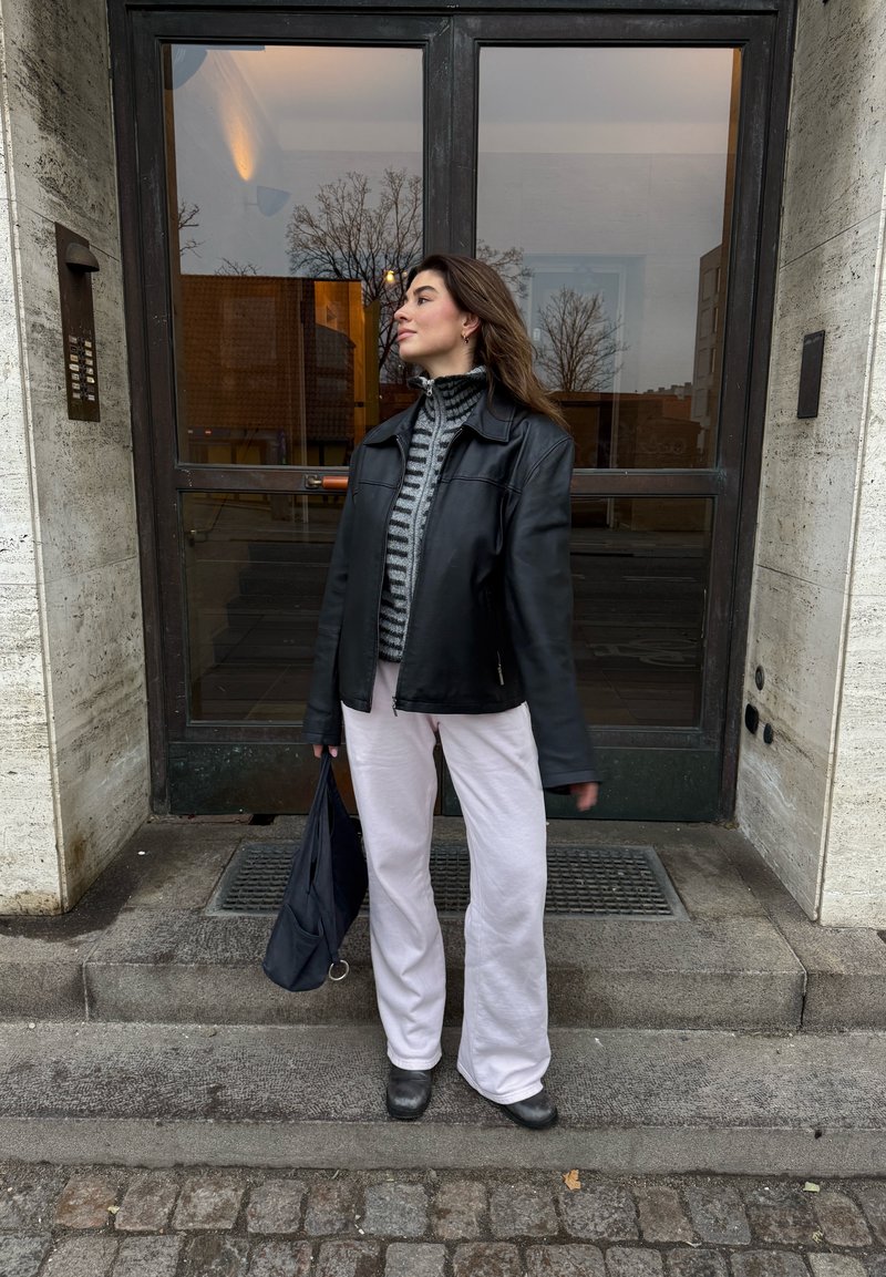 Veste en cuir noire portée sur un pull à col montant rayé, associée à un pantalon large rose clair et des chaussures noires, tenant un sac sombre.
