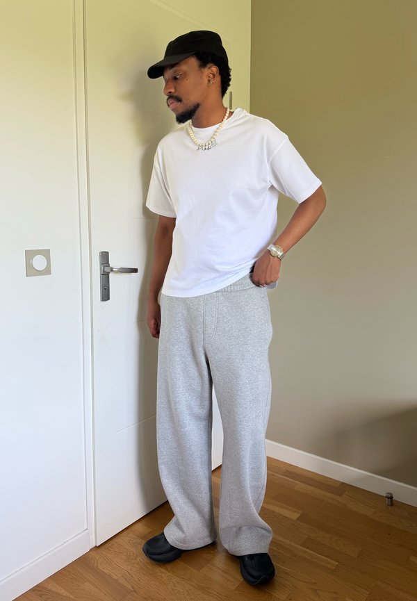 Mand iført sort kasket, hvid t-shirt, grå joggingbukser, sorte sko og en halskæde, der står indenfor ved en lukket hvid dør.