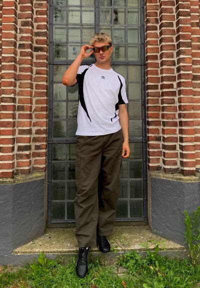 Camisa atlética blanca con detalles en negro, pantalones cargo verde oliva y zapatos negros. De pie junto a una pared de ladrillo, ajustando unas gafas de sol con tintado naranja.