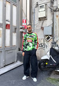 Άνδρας που φοράει φωτεινό πράσινο γραφικό φούτερ και μαύρο παντελόνι cargo στέκεται δίπλα σε σκούτερ και βιομηχανικό κτίριο με προειδοποιητικές πινακίδες και γκράφιτι.