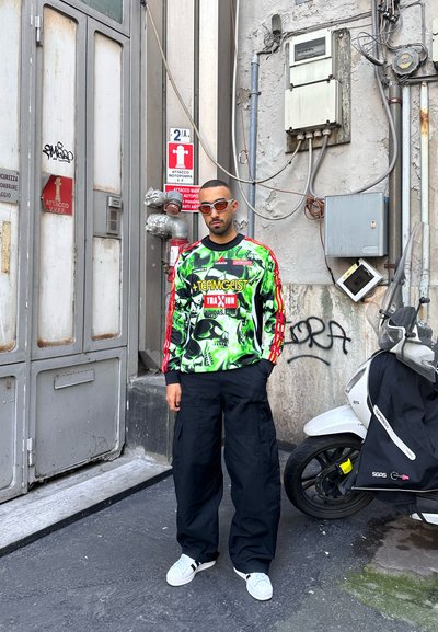 Hombre con sudadera gráfica verde brillante y pantalones cargo negros de pie junto a un scooter y un edificio industrial con señales de advertencia y grafitis.