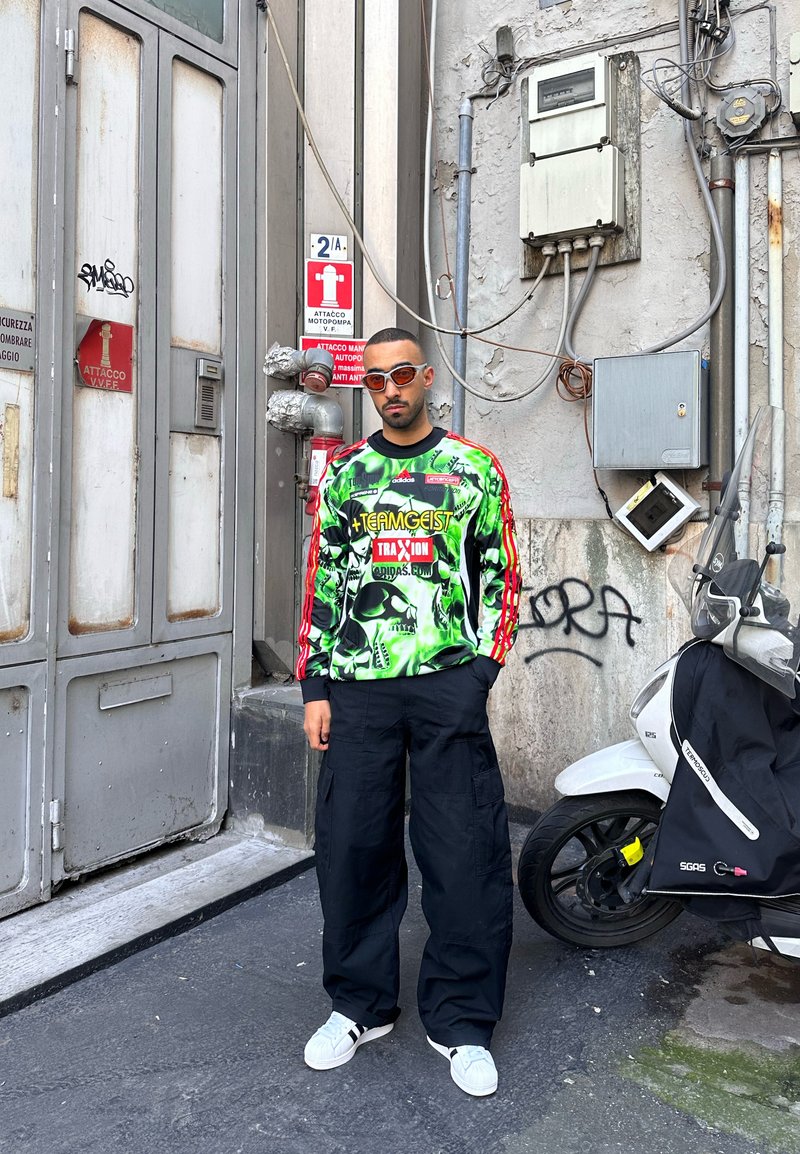 Uomo con felpa grafica verde brillante e pantaloni cargo neri in piedi accanto a un monopattino e a un edificio industriale con cartelli di avviso e graffiti.