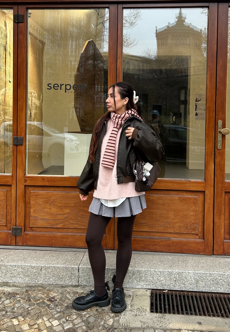 Young woman with long hair stands outside a gallery, wearing a leather jacket, striped scarf, pleated skirt, and carrying a handbag.