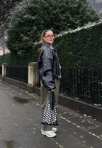 Chaqueta corta de cuero negra, pantalones verde oliva, zapatillas gris, sosteniendo un bolso negro con tachuelas plateadas, de pie frente a un fondo de seto.