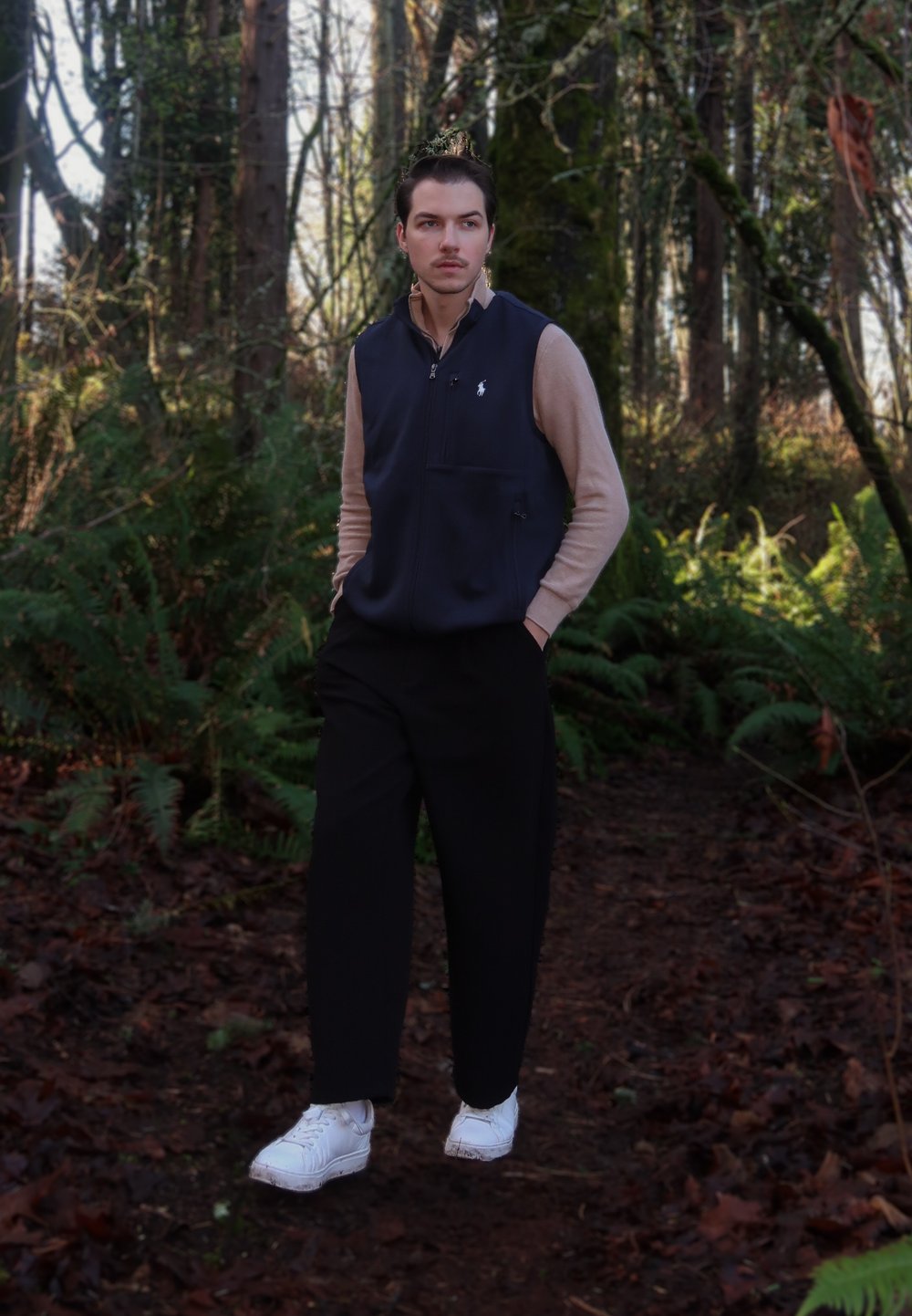 Veste zippée bleu marine portée sur une chemise beige à manches longues, accompagnée d'un pantalon noir et de baskets blanches, dans un environnement forestier avec du feuillage.