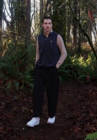 Veste zippée bleu marine portée sur une chemise beige à manches longues, accompagnée d'un pantalon noir et de baskets blanches, dans un environnement forestier avec du feuillage.