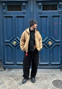 Tan hooded jacket worn over a black hoodie, paired with black wide-leg trousers and black shoes. Wearing a grey patterned beanie. Standing in front of blue doors.