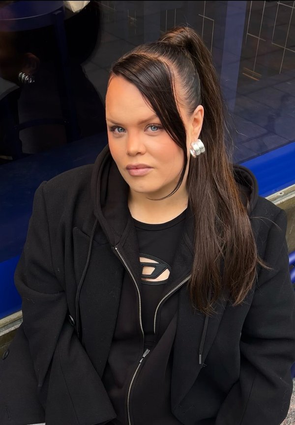 Zwarte hoodie met rits over een zwart topje met een uitsnijding. Lang donker haar in een hoge paardenstaart en zilveren cirkelvormige oorbellen. Blauwe achtergrond.