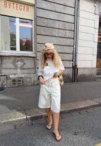 Vit bomull t-shirt, beige denim-shorts, färgglad mönstrad huvudduk, guldiga solglasögon och bruna flip-flops; står på en stadsgata.