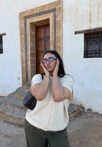 Personne aux longs cheveux noirs et lunettes, posant avec les mains sur les joues devant des marches en pierre et une porte en bois encadrée de carreaux beige et bleu.