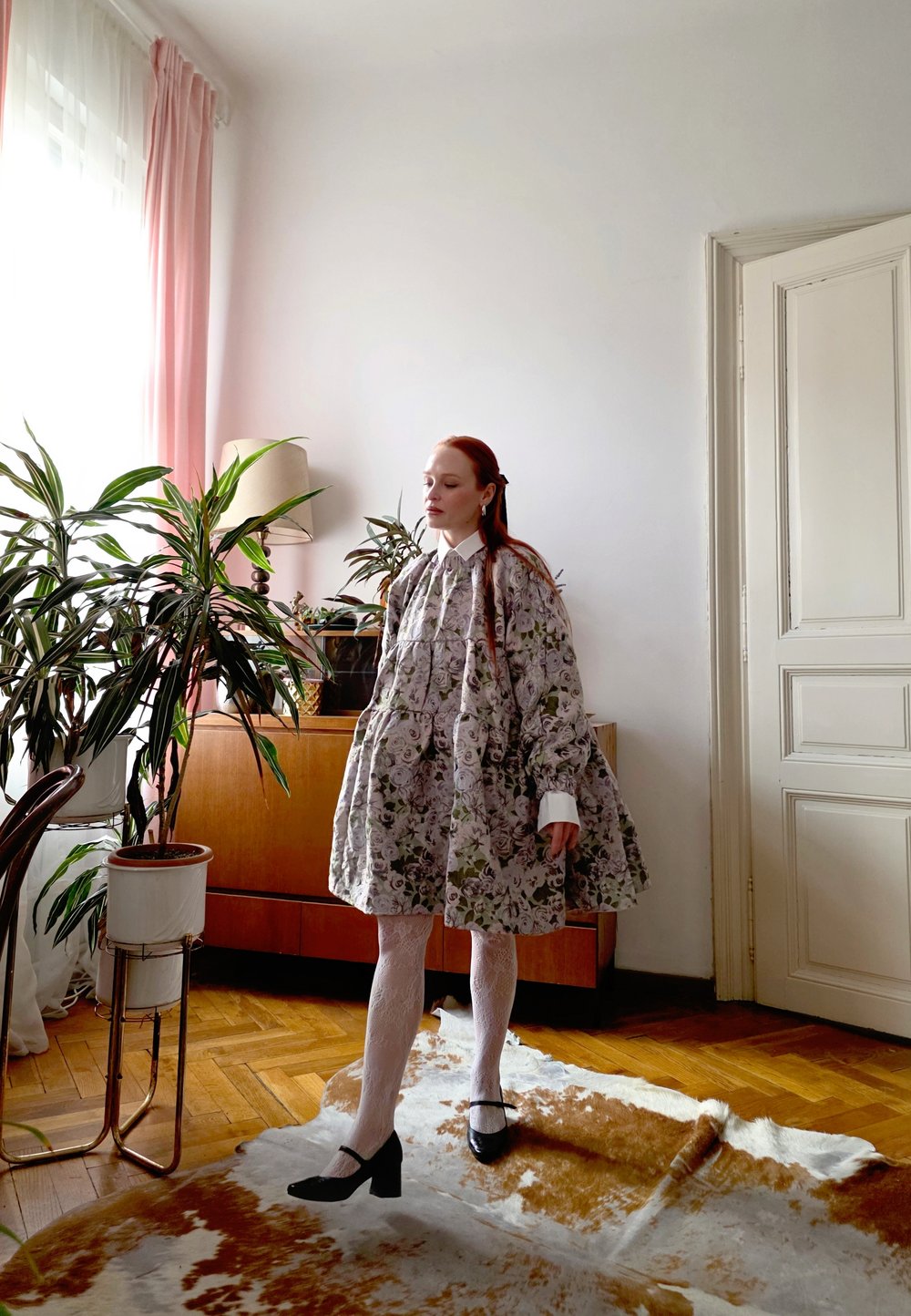 Kleita ar ziedu rakstu gaišā audumā, ar vairākām kārtām un baltām aprocēm. Modelis valkā baltas zeķubikses un melnus papēžus. Telpa ar koka mēbelēm.