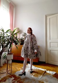 Kleita ar ziedu rakstu gaišā audumā, ar vairākām kārtām un baltām aprocēm. Modelis valkā baltas zeķubikses un melnus papēžus. Telpa ar koka mēbelēm.