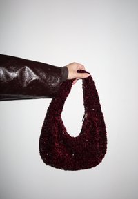 Main tenant un petit sac à main en velours rouge foncé texturé avec une courte bandoulière sur un fond blanc uni.