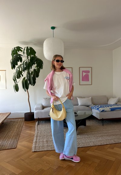 Pantalones de mezclilla anchos de color claro, camiseta blanca, cárdigan rosa caído sobre los hombros, bolso amarillo y zapatillas rosas. Entorno interior con plantas.
