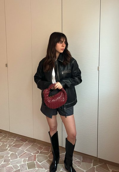 Chaqueta de cuero negra sobre una camisa blanca, combinada con shorts grises y botas negras hasta la rodilla. Sosteniendo un bolso de mano trenzado rojo.