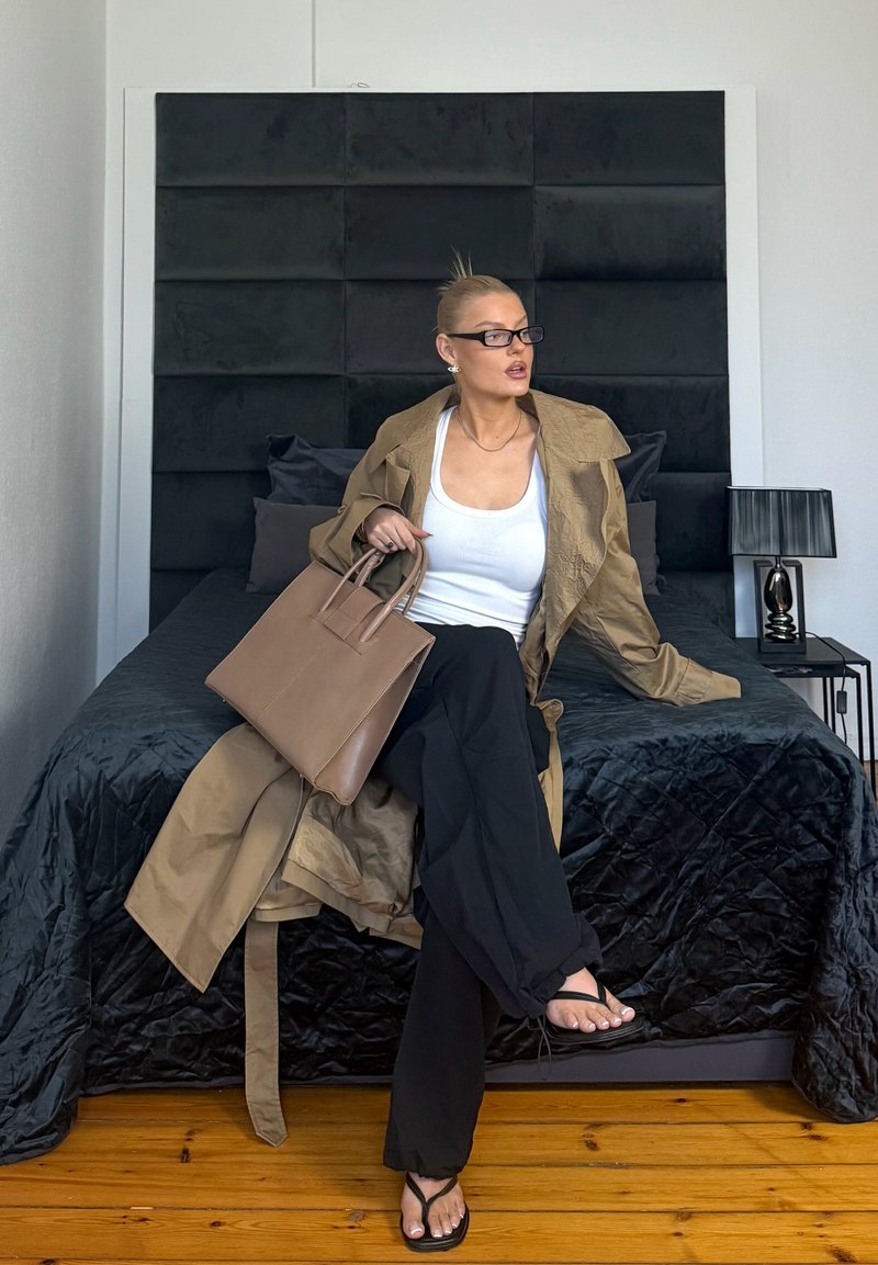 Prillidega naine, kes kannab beežikat trenkjope, valget toppi ja musti pükse, istub voodil, hoides käes beežikat käekotti moodsas magamistoas.