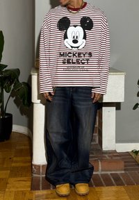 Raudonos ir baltos juostelėmis puošta ilgomis rankovėmis marškinėliai su dideliu Mikio Pelytės piešiniu ir užrašu, derinami su laisvo kirpimo tamsaus džinso džinsais ir smėlio spalvos klumpėmis.