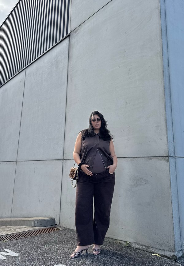 Mujer con camiseta sin mangas oscura y pantalones anchos, gafas de sol y zapatos con estampado de leopardo, está de pie junto a una alta pared de hormigón al aire libre.