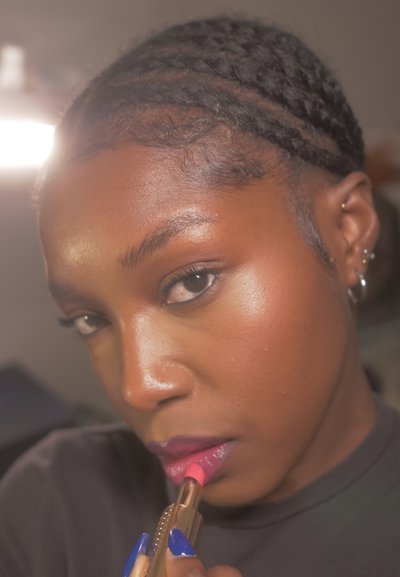 Primer plano del rostro de una persona con cabello negro trenzado, aplicándose un brillo labial color rosa brillante desde un tubo dorado, mostrando una textura labial brillante.