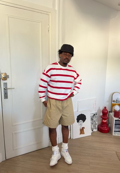 Hombre con pantalones cortos beige, camisa de manga larga a rayas rojas y blancas, calcetines blancos, zapatillas y gorra negra de pie cerca de una puerta blanca en una habitación con arte y monopatín.
