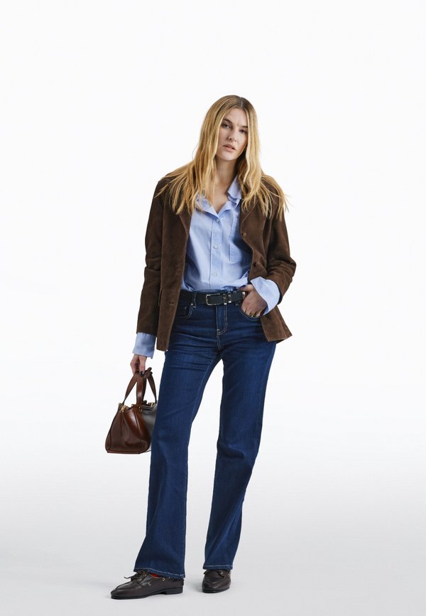 Γυναίκα που στέκεται φορώντας καφέ μπουφάν, ανοιχτό μπλε πουκάμισο, σκουρό μπλε τζιν με καμπάνα, μαύρα παπούτσια και κρατώντας καφέ τσάντα στο αριστερό της χέρι.