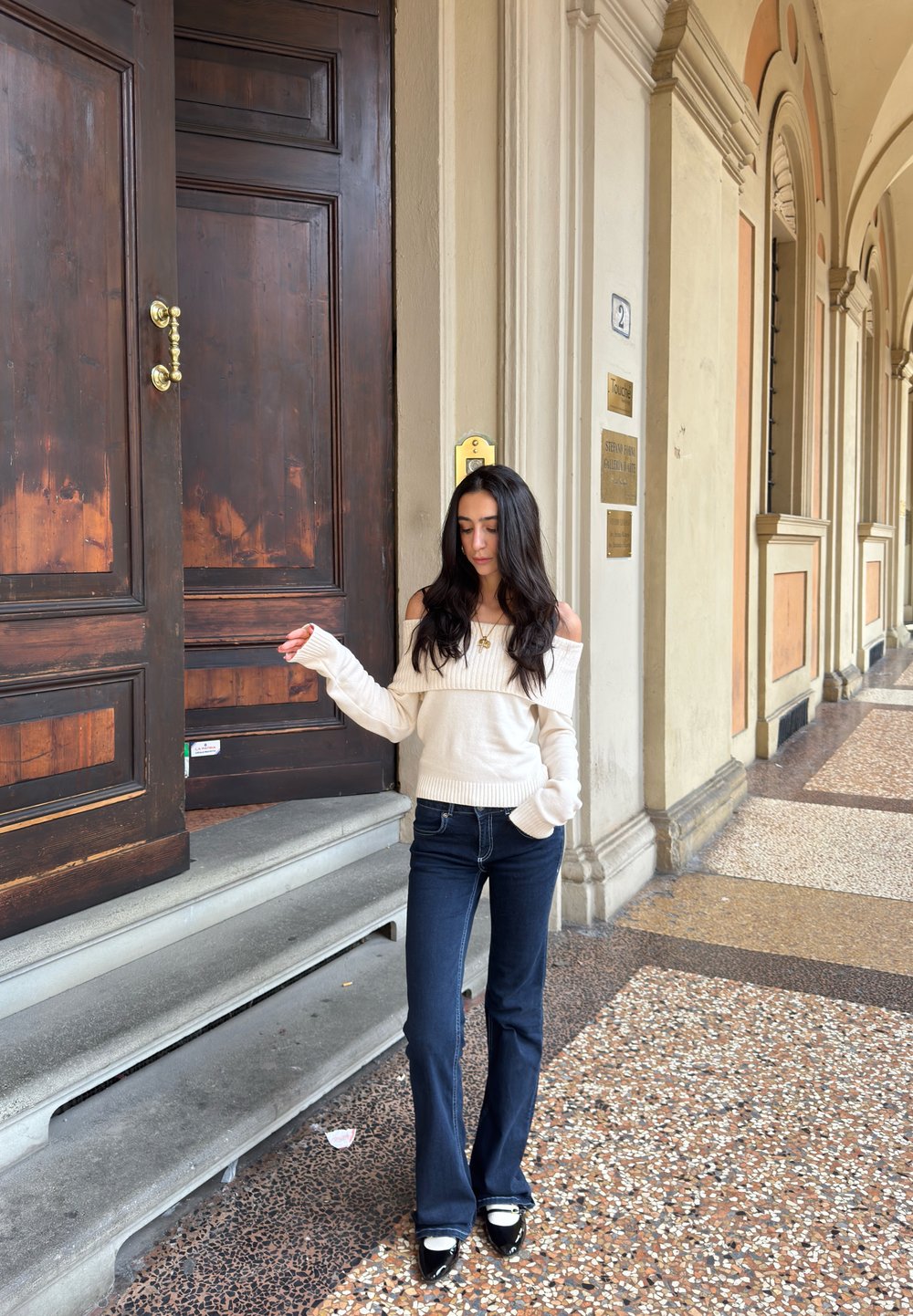 Pull en tricot crème décolleté avec une texture côtelée, associé à un jean flare sombre. Avec des chaussures à talons noires et blanches, se tenant près de portes en bois.
