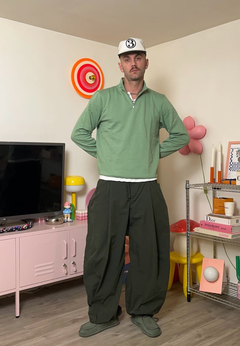 Maglione verde con zip, pantaloni oversize verde scuro e scarpe verdi abbinate. T-shirt bianca sotto, cappellino con logo circolare.
