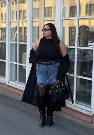 Mujer con top negro sin mangas, falda vaquera, medias de encaje, botas negras, sosteniendo bolso acolchado, de pie junto a grandes ventanas de cristal.