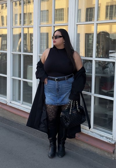 Mujer con top negro sin mangas, falda vaquera, medias de encaje, botas negras, sosteniendo bolso acolchado, de pie junto a grandes ventanas de cristal.