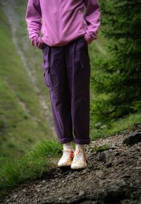 Paarse cargo broek met zijzakken, gecombineerd met witte sportschoenen met rode accenten. Zachte roze fleecejack, staande op een rotsachtige pad.