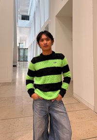 Mies, jolla on vihreä-mustaraidallinen pörröinen villapaita ja leveälahkeiset farkut, seisoo käsivartensa taskuissa kirkkaassa modernissa käytävässä.