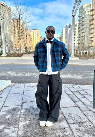 Chaqueta a cuadros en azul y negro con dos bolsillos en el pecho, sobre una camiseta blanca, combinada con pantalones cargo negros holgados y zapatillas blancas.