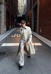 Béžový trenčkot s golierom na svetlošedý outfit, doplnený čiernymi topánkami a slnečnými okuliarmi, kráča v úzkej mestskom uličke.